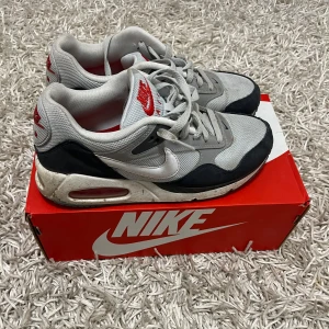 Nike Air Max  - Säljer ett par klassiska Nike Air Max sneakers i grått och svart med röda detaljer. Skorna har en synlig luftkudde i sulan för extra komfort och snörning framtill. Perfekta för både vardag och träning. De är i bra skick med lite slitage på sulan.