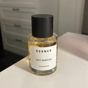 Essnce Hot Mimosa Eau de Parfum - Säljer en elegant flaska Essnce Hot Mimosa Eau de Parfum. Flaskan är rund och genomskinlig med en stilren svart kork. Doften är fräsch och blommig, perfekt för både vardag och fest. En lyxig touch till din parfymkollektion!