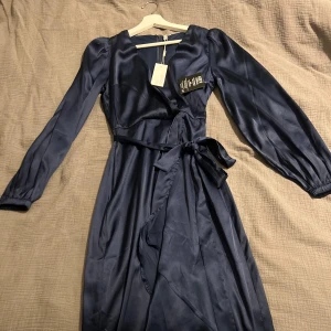 Mörkblå omlottklänning - Säljer en elegant mörkblå omlottklänning i siden med långa ärmar och knytband i midjan. Perfekt för festliga tillfällen eller en fin middag. Klänningen har en v-ringad hals och ger en lyxig känsla. Passar bra till både höst och vinter. Klänningen kommer ifrån NELLY.COM och är helt ny med alla lappar kvar. (Köparen står för frakt) 