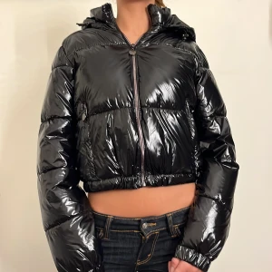 Svart glansig pufferjacka - Säljer en supercool svart glansig pufferjacka med dragkedja. Den är croppad och har en luva, perfekt för att hålla värmen. Jackan är i mycket bra skick och passar perfekt för höst och vinter. Den har långa ärmar och en snygg, modern look.