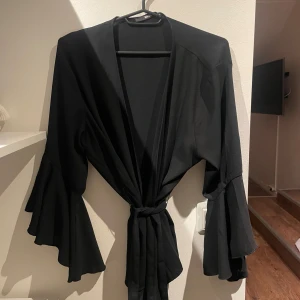 Svart kimono från Bik Bok - Säljer nu den här blusen från Bik Bok i storlek M! Den har långa ärmar med volangdetaljer och ett knytband i midjan❣️
