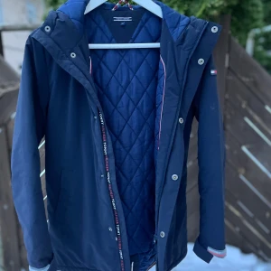 Mörkblå jacka från Tommy Hilfiger - Säljer en snygg mörkblå jacka från Tommy Hilfiger. Den har en quiltad insida och en huva för extra skydd mot väder. Jackan har dragkedja och knappar framtill, samt ett stort Hilfiger-tryck på ryggen. Perfekt för höst, vinter och vår!