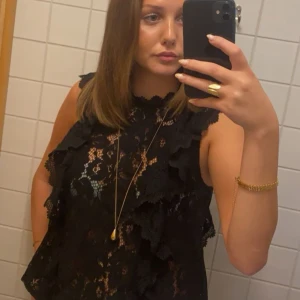 Svart spetsblus - Säljer en elegant svart spetsblus med volangdetaljer. Den är ärmlös och perfekt för festliga tillfällen men också bara till skolan. Blusen har en fin passform och är gjord i ett mjukt spetsmaterial som ger en lyxig känsla.