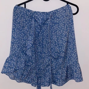 Blå blommig kjol - Söt blå blommig kjol med volangdetaljer. Perfekt för sommardagar eller en avslappnad vårlook. Kjolen har en omlott-design som ger en fin passform och rörelse. Materialet känns lätt och luftigt, vilket gör den idealisk för varmare väder.