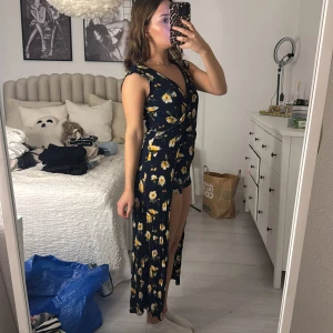 Blommig omlottklänning - Säljer en superfin blommig omlottklänning i mörkblått med gula och vita blommor. Klänningen har en v-ringad design och en slits som ger en snygg look. Perfekt för sommarens fester eller en dag på stan. Den är i mycket bra skick och passar perfekt för en avslappnad men stilren outfit.