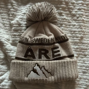 Åre mössa! - Säljer en mysig beige stickad mössa med stor tofs. Den har texten 'Åre' och ett bergsmotiv broderat framtill. Perfekt för kalla vinterdagar i skidbacken eller på stan. Ribbad kant för extra värme och stil.