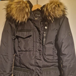 Svart Livigno från Hollies med äkta päls strl 34 - Säljer en snygg svart och varm vinterjacka Livigno från Hollies i storlek 34. Jackan har en fluffig äkta pälsfodrad huva och flera praktiska fickor med både knappar och dragkedja. 🖤