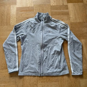 Grå träningsjacka från Adidas - Säljer en snygg grå träningsjacka från Adidas i bra skick. Jackan har dragkedja och långa ärmar med vita detaljer längs sidorna. Perfekt för träning eller som en casual vardagslook. Den är gjord i mjukt bomullsmaterial och har en normal passform.