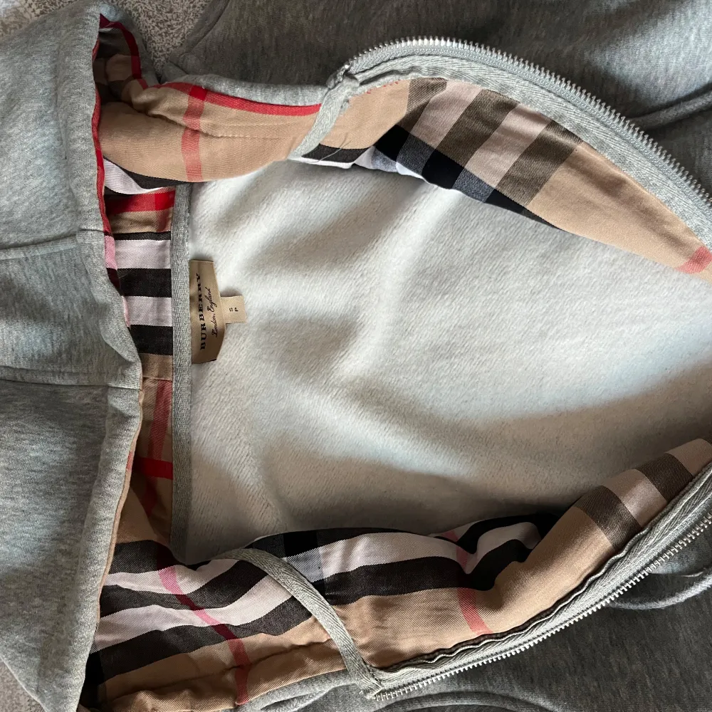 Säljer en stilren grå hoodie från Burberry i mycket bra skick. Den har en dragkedja framtill och en klassisk Burberry-rutig insida i huvan. Perfekt för höst och vår, med en normal passform och långa ärmar. Superbekväm och snygg för vardagsbruk!. Tröjor & Koftor.