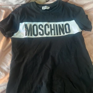 Svart t-shirt från Moschino - Säljer en svart t-shirt från Moschino med stort vitt tryck på bröstet. Den är i bra skick och perfekt för en casual look. Passar både till vardags och fest. Storleken är för tonåringar.