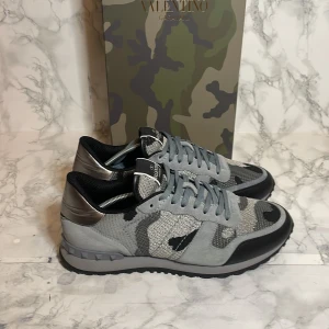 Valentino Rockrunner - Säljer ett par snygga Valentino Rockrunner sneakers i grått och svart med coolt camouflage-mönster. Skorna är i mycket bra skick och har en unik design med nitar på hälen. Perfekta för vardagen