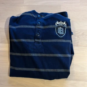 Mörkblå randig tröja från Bondelid - Säljer en snygg mörkblå tröja från Bondelid med grå ränder och ett stiligt emblem på bröstet. Tröjan har knappar framtill och en cool patch på ärmen. Perfekt för en avslappnad men stilren look! Fint skick men passar ej längre!