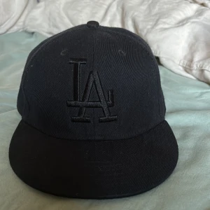 Svart LA keps storlek 55 cm - Snygg svart keps med broderad LA-logga fram och bak. Kepsen är i bra skick och passar de flesta med sin storlek på 55 cm. Hör av dig vid intresse!