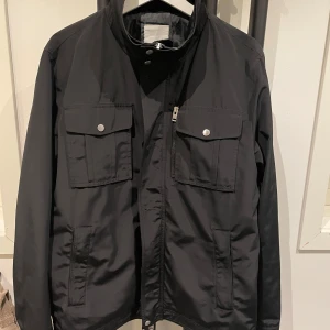 Svart jacka från J.Lindeberg - Säljer en stilren svart jacka från J.Lindeberg i storlek L. Jackan har en dragkedja och knappar framtill, samt praktiska fickor med lock. Perfekt för höst och vår, med en modern och tidlös design. Passar både till vardags och lite finare tillfällen. 