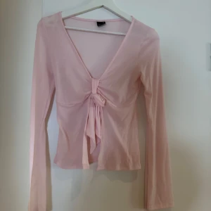 Rosa långärmad topp med knyt - Säljer en söt rosa långärmad topp med knyt framtill. Den har en v-ringad design och är perfekt för både vardag och fest. Toppen är i ett mjukt och bekvämt material som känns skönt mot huden.