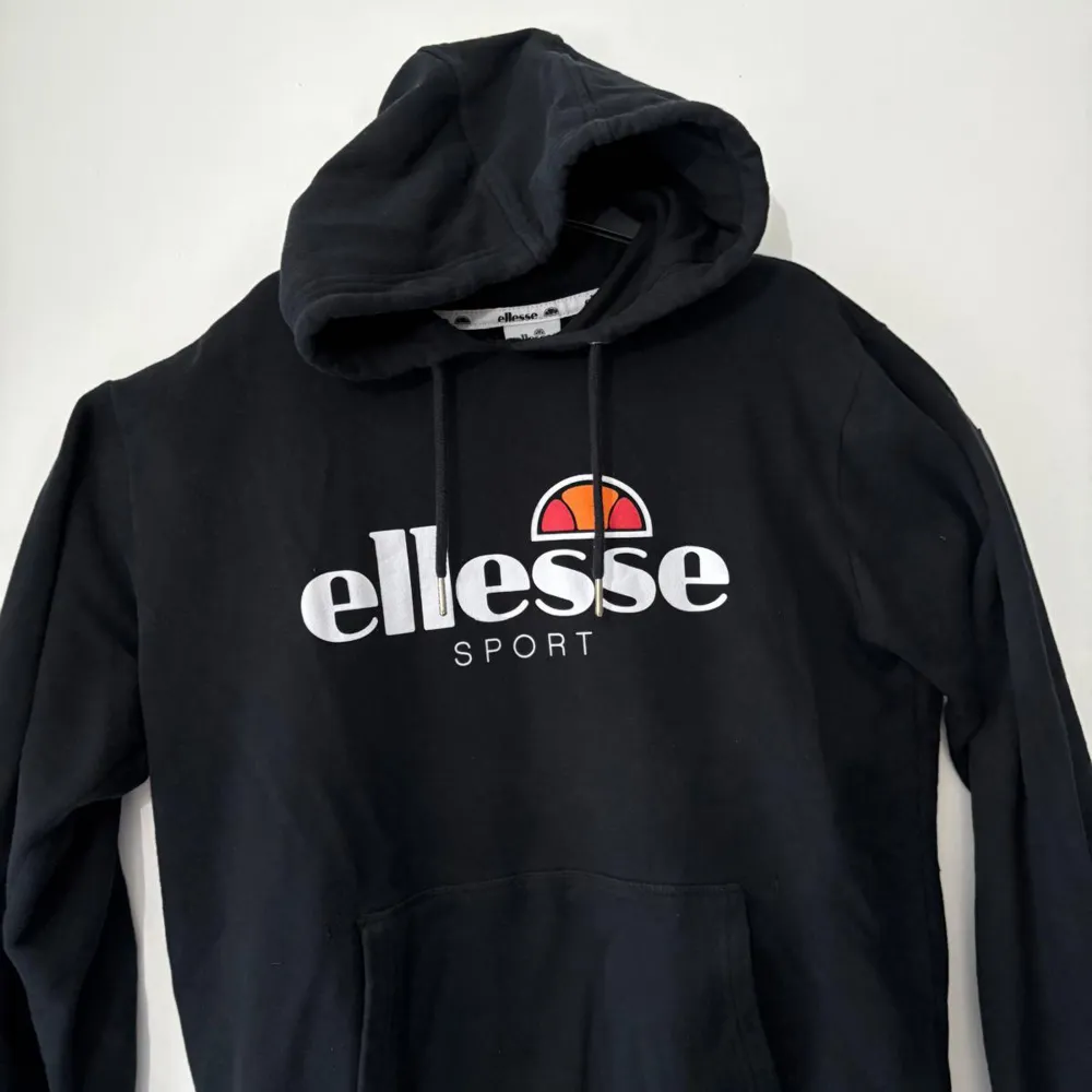 Säljer en snygg svart hoodie från Ellesse med deras ikoniska logga i vitt och orange på bröstet. Perfekt för en sportig look eller bara för att chilla. Den har en klassisk känguruficka och justerbar huva med dragsko. Passar perfekt till höst och vinter!. Tröjor & Koftor.