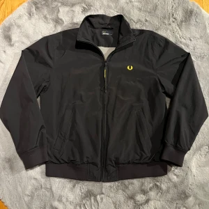 Svart jacka från Fred Perry - Säljer en snygg svart jacka från Fred Perry i storlek M. Jackan har en dragkedja framtill och det klassiska Fred Perry-emblemet broderat i gult på bröstet. Perfekt för höst och vinter med sitt stilrena och tidlösa utseende. Innerfodret är i ett meshmaterial för extra komfort.