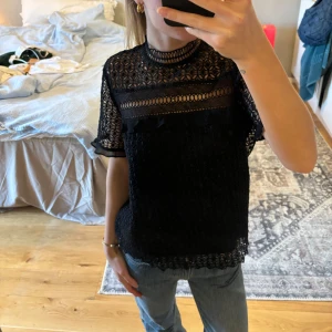 Svart spetsblus - Säljer en elegant svart spetsblus med korta ärmar och hög hals. Blusen har ett vackert mönster och är perfekt för en festlig kväll. Den är lätt och luftig, vilket gör den bekväm att bära. Passar bra till både jeans och kjol för en chic look.