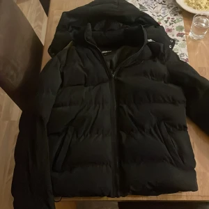 Svart pufferjacka från Urban Classics - Säljer en svart pufferjacka från Urban Classics i storlek s.Jackan är perfekt för kalla dagar med sin vadderade design och har en praktisk dragkedja framtill. Den är i mycket bra skick och passar både höst och vinter. Huvan ger extra skydd mot kyla. Perfekt för dig som vill hålla stilen i kylan! 🖤