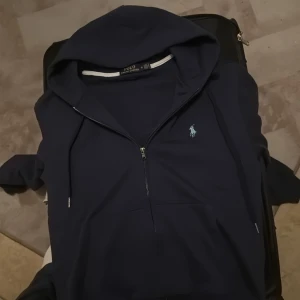 Mörkblå hoodie från Ralph Lauren - Säljer en snygg mörkblå hoodie från Ralph Lauren i storlek M. Den har en dragkedja framtill och en klassisk logga på bröstet. Perfekt för en avslappnad stil och passar bra till både höst och vår. Hör av dig vid intresse!