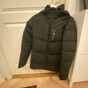 Svart pufferjacka från Sail Racing - Säljer en svart pufferjacka från Sail Racing gammal och pyttelite sliten men användbar, varm och stilig! Jackan har en dragkedja framtill och en praktisk huva. Perfekt för kalla vinterdagar med sin varma och vadderade design. Den har också en snygg logga på bröstet och ärmen. Passar både till vardags och utomhusaktiviteter. Storlek 170 köpt för 3000 i kidsbrandstore för ungefär 2 år sedan.