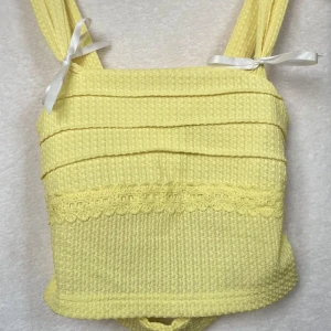 Gul ribbad bikini med spetsdetaljer - Superfin gul topp i ribbat material med söta spetsdetaljer. Den har breda axelband med vita rosetter och en knytning i ryggen för en perfekt passform.💛
