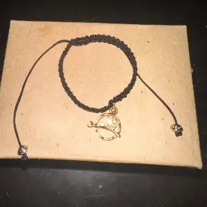 Säljer ett snyggt svart armband med en guldberlock som föreställer en katt på en pinne. Armbandet är flätat och justerbart, perfekt för att ge en unik touch till din stil. Passar både till vardags och fest!