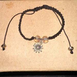 Svart armband med blomma och pärlor - Säljer ett snyggt svart armband med en justerbar snörning. Armbandet har två genomskinliga pärlor och ett hängsmycke i form av en blomma i silverfärg. Perfekt för att ge en bohemisk touch till din outfit!