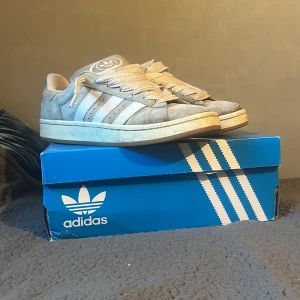 Grå Adidas Campus 00s - Säljer ett par gråa Adidas Campus 00s sneakers i storlek herr 43. Skorna är i en snygg grå färg med vita detaljer och klassiska tre ränder på sidan. De är i bra skick och kommer med originalkartong. Perfekta för både vardag och fest!