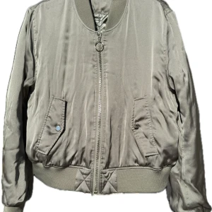 Olivgrön bomberjacka - Olivgrön bomberjacka med dragkedja framtill. Jackan har långa ärmar och ribbade muddar vid ärmslut och nederkant. Perfekt för höst och vår! Ca 38/40 till dam eller small till kille. Hämtas på Hägerstensåsen eller ök 💚