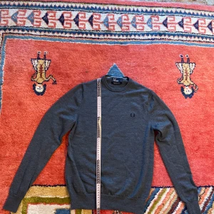 Grå ulltröja från Fred Perry -  grå tröja från Fred Perry i 100% merinoull. Inga fläckar samt slitage förekommer. Måtten är följande:   Längd, ifrån toppen av axeln, ca 61 cm    bredd, armhåla till armhåla, ca 44cm  armlängd, ifrån armhåla utot, ca 48cm ärm öppning, ca 6cm
