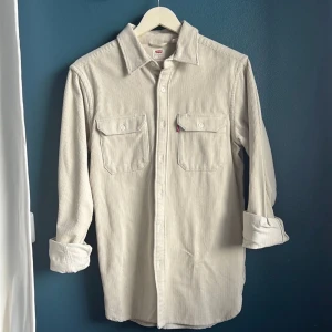 Levi’s Manchesterskjorta, avslappnad passform  - En skjorta med långa ärmar och två fickor på bröstet. Den har en knappslå på framsidan och knappar vid ärmsluten. Obs, en knapp har lossnat vid ärmslutet, finns extra att sy fast om man vill kavla ner ärmarna.