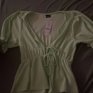 Grön Daisy topp - Säljer en superfin ljusgrön topp i storlek XS. Den har en v-ringad design med knytning framtill och puffiga korta ärmar. Perfekt för vår och sommar! 💚