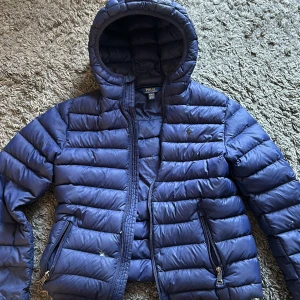 Blå dunjacka från Ralph Lauren - Säljer en snygg blå dunjacka från Ralph Lauren. Jackan är quiltad och har en praktisk huva. Den är perfekt för kyliga dagar och har dragkedja framtill samt fickor med dragkedjor. Jackan är i storlek M och är i bra skick.