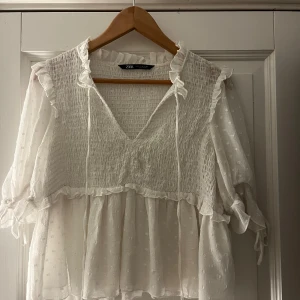 Vit blus från Zara - Säljer en superfin vit blus från Zara. Den har en v-ringad design med volangdetaljer och trekvartsärmar. Blusen är gjord i ett lätt och luftigt material, perfekt för vår och sommar. Den har också en söt knytning vid ärmsluten.