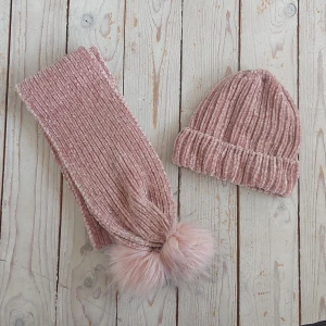 Rosa stickad mössa och halsduk med pälstofs - Superfin rosa stickad mössa och matchande halsduk. Mössan är ribbad och ser riktigt mysig ut för kalla dagar. Halsduken har fluffiga pälstofsar i änden som ger en extra touch av stil. Perfekt för att hålla sig varm och snygg under vintern!