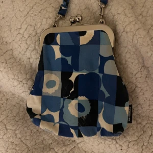 Blå mönstrad väska från Marimekko - Säljer en superfin blå mönstrad väska från Marimekko. Den har ett coolt retroinspirerat mönster i olika blå nyanser och vitt. Väskan har en metallram med en klassisk bygelstängning och en kort axelrem. Perfekt för att ge en färgklick till din outfit!