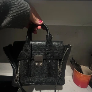Svart skinnväska med dragkedja - Säljer en snygg svart skinnväska med coola detaljer. Väskan har en praktisk dragkedja och ett stilrent metallspänne framtill. Perfekt för både vardag och fest! Från Phillip Lim