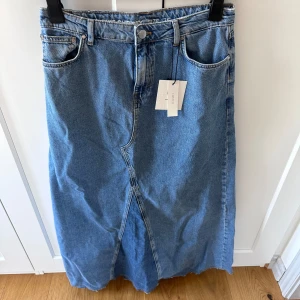 Maxikjol i jeans - CHRISTY MAXIKJOL JEANS - BikBok Never Denim En jeanskjol i 100% återvunnen bomull. Denimkjolen har låg midja med kontrastpaneler på framsidan, stängs med gylf, knapp och hällor. Kjolen har en hög slit bak.  Är aldrig använd - har alla lappar kvar.