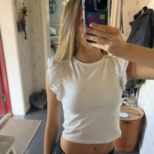 Vit croppad topp - Säljer en superfin vit croppad topp med volangdetaljer på ärmarna. Toppen är tight och ärmlös, vilket gör den både bekväm och fin! Den är i nyskick och endast använd 1 gång🥰