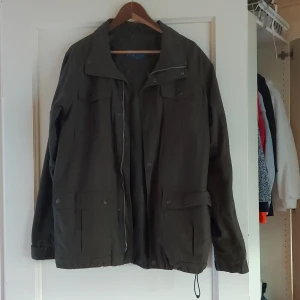 Grön vindjacka - Säljer en grön vindjacka som är perfekt för höst och vår. Jackan har en dragkedja framtill och flera praktiska fickor med knappar. Den är långärmad och har en justerbar dragsko i nederkanten för bättre passform.