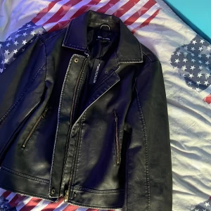 Svart skinnjacka från PrettyLittleThing - Säljer en snygg svart skinnjacka från PrettyLittleThing. Jackan har en cool bikerstil med dragkedja och knappar. Perfekt för höst och vår, och den funkar lika bra till fest som till vardags. Den är i nyskick.