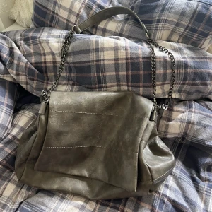 Grå axelväska med kedjedetaljer - Säljer en snygg grå axelväska i skinnimitation med coola kedjedetaljer. Väskan har en praktisk dragkedja och är perfekt för både vardag och fest. Den är rymlig och har en stilren design som passar till det mesta.