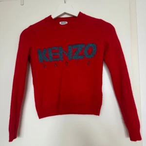 Röd ulltröja från Kenzo - Säljer en superfin röd tröja från Kenzo i storlek S (men är snarare Xs). Den är gjord av 96% ull, vilket gör den perfekt för kyliga dagar. Tröjan har långa ärmar och ett stort Kenzo Paris-tryck på framsidan. Den är både snygg och bekväm, perfekt för höst och vinter!