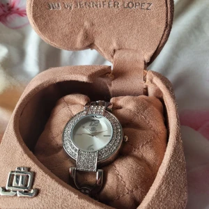 Glittrig klocka från JLO by Jennifer Lopez - Säljer en superfin och glittrig klocka från JLO by Jennifer Lopez. Klockan har en rund urtavla med silverfärgade detaljer och är dekorerad med gnistrande stenar runt om. Armbandet är i silver och har en elegant länkdesign med fler glittrande stenar. Perfekt för festliga tillfällen! Kommer i en snygg, beige förvaringsbox.