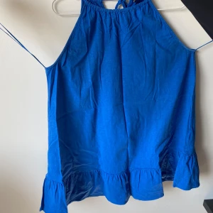 Blå klänning från Zara - Säljer en superfin blå klänning från Zara. Den är ärmlös och har en volangdetalj längst ner. Perfekt för sommaren med sin lätta och luftiga stil. Klänningen har en knytning i nacken som ger en söt touch. Passar perfekt till både vardag och fest!