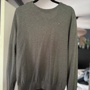 Grå tröja från Tommy Hilfiger - Säljer en stilren grå tröja från Tommy Hilfiger. Den är i storlek M och har en liten broderad logga på bröstet. Perfekt för både vardag och lite finare tillfällen. Tröjan är i bra skick och superbekväm!
