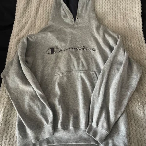 Grå hoodie från Champion - Säljer en grå hoodie från Champion. Den har en Championlogga på bröstet och en liten logga på ärmen. Hoodien är långärmad och har en stor ficka framtill. Den är i bomullsmaterial och har en liten slitage vid mudden. Kontakta mig om du har fler frågor 😊