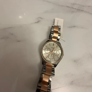 Silver och roséguld klocka från Regal - Oanvänd klocka från Regal i silver och roséguld med en glittrig kant runt urtavlan. Klockan har tre små urtavlor och en stilren design som passar perfekt för den som vill ha något extra på handleden.