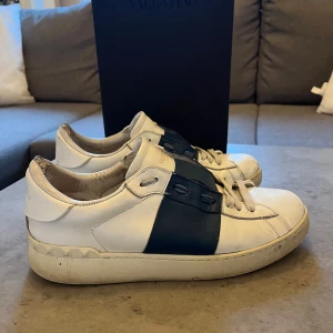 Valentino sneakers i vitt och blått - Snygga Valentino sneakers i vitt med en bred blå rem över snörningen. Skorna har en klassisk design med rund tå och platt sula. Perfekta för en stilren look. Finns defekter vid hälen och ganska slitna under skon.